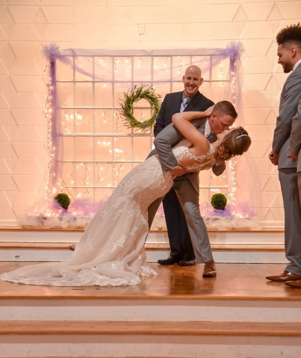 Groom dips bride during wedding ceremony; both smile. Decorated backdrop, two attendants.