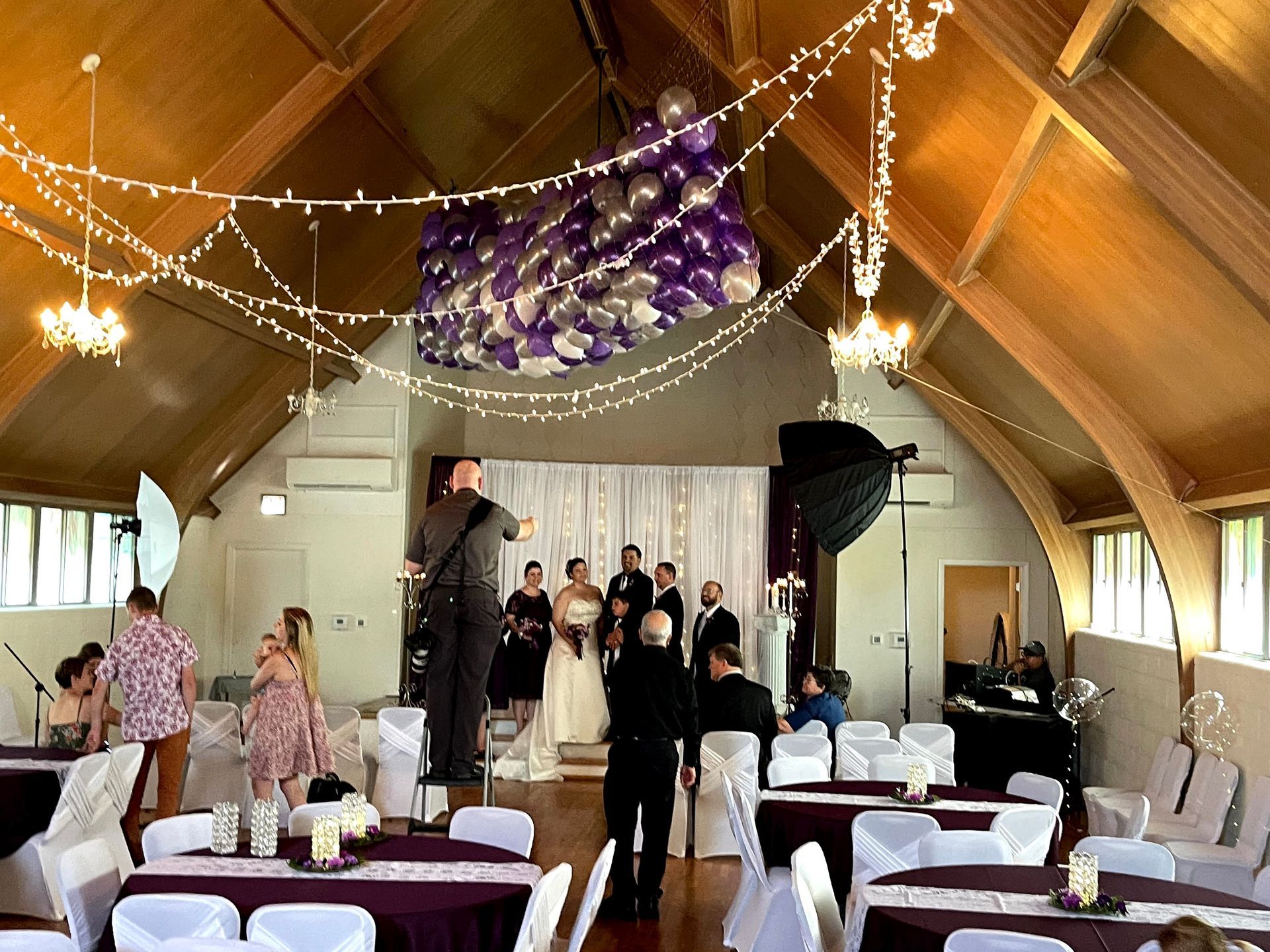 Wedding reception in a hall with a photographer taking a photo of the wedding party. Tables are set with purple and white decor.