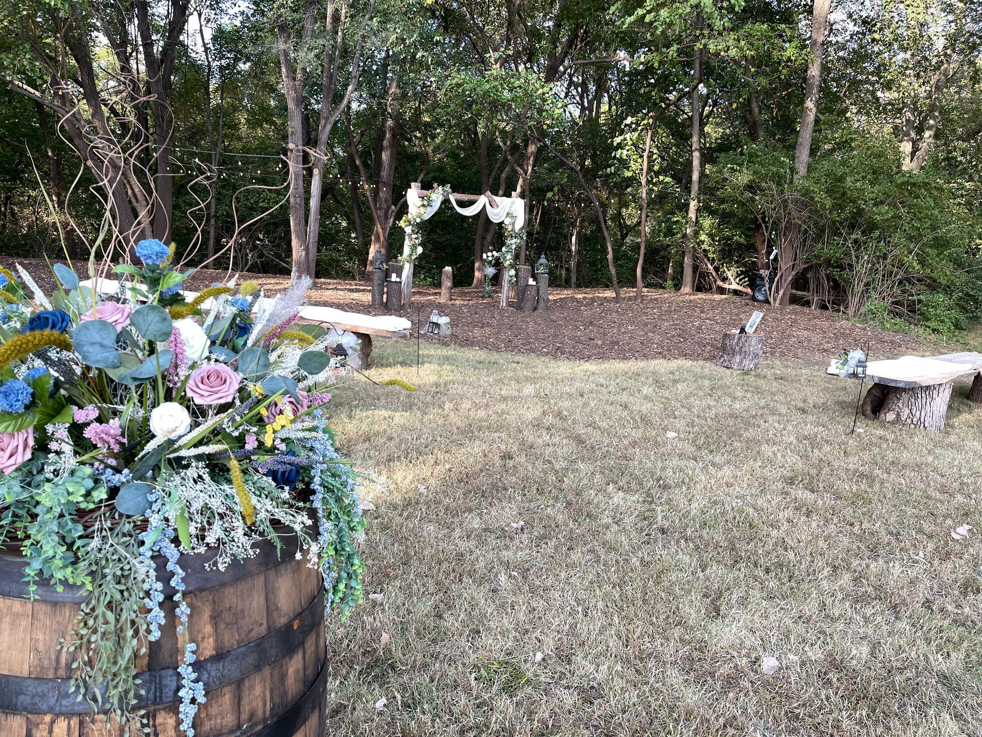 Wedding ceremony setup in a wooded area; floral arrangement on a barrel in foreground, arch in the background.