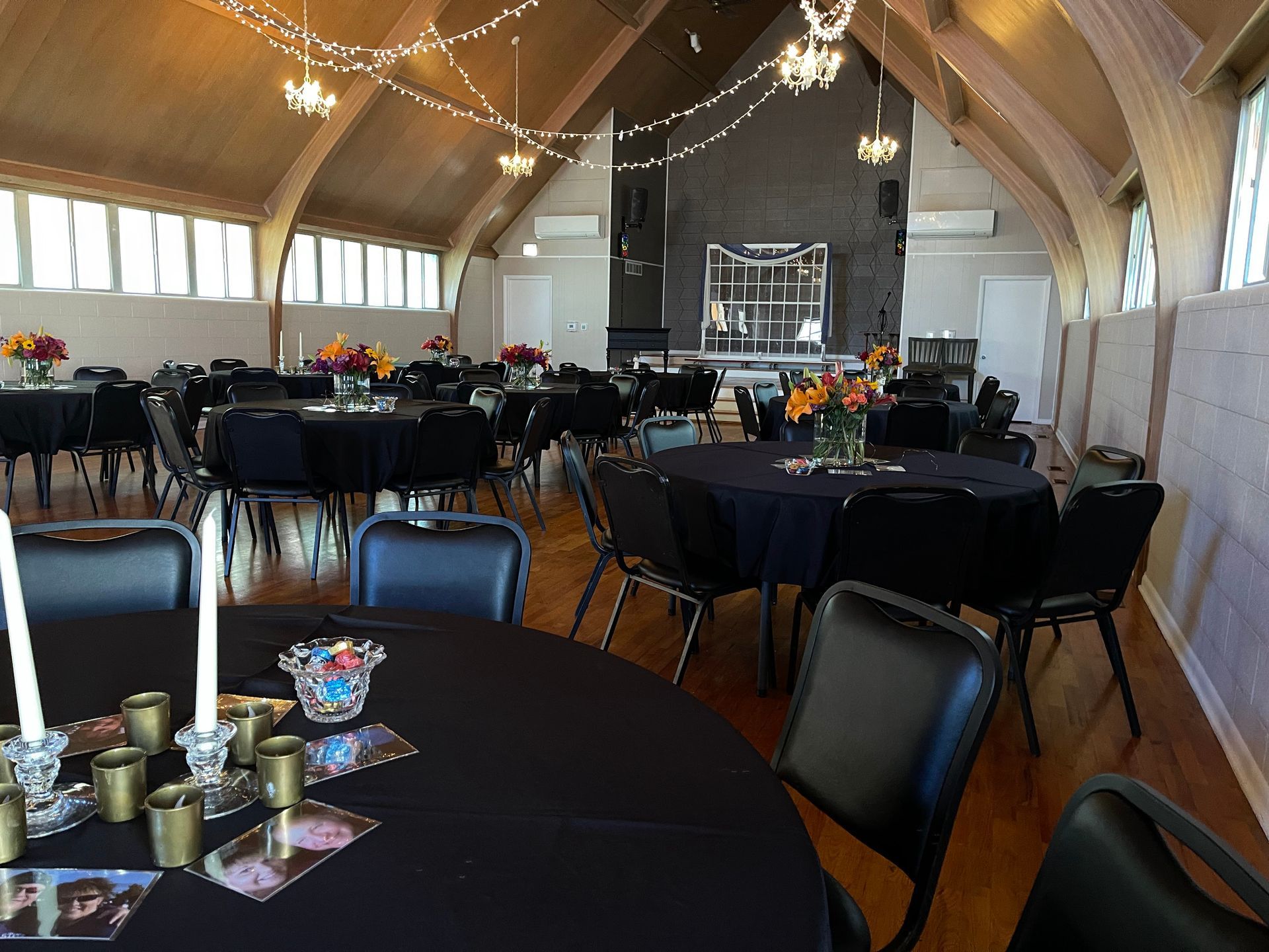 Event hall set with round tables, black linens, and flowers. Fairy lights hang from the high arched ceiling.