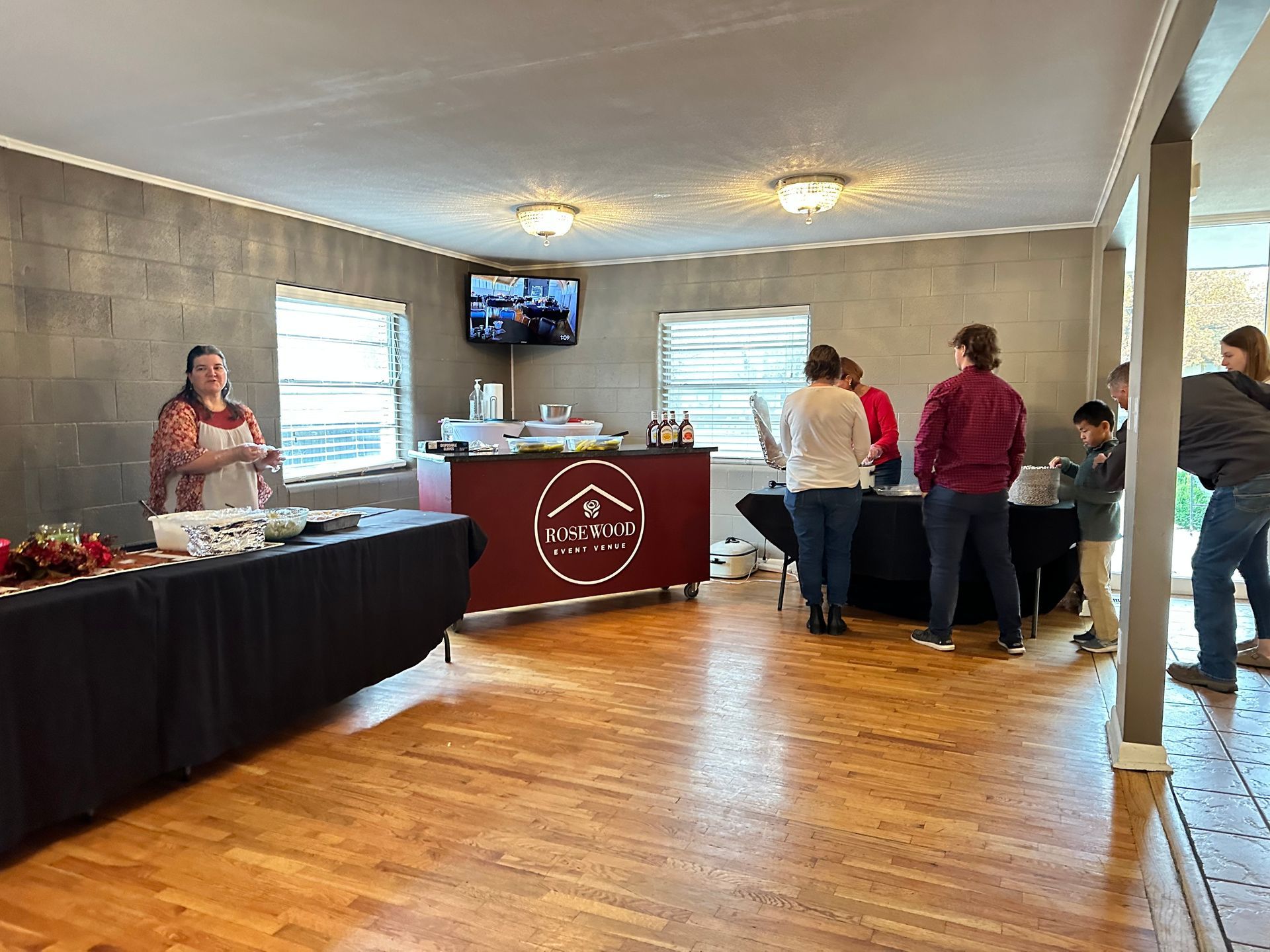 People at a food tasting event. Tables with samples, signage, and people in a room with windows and wood floors.