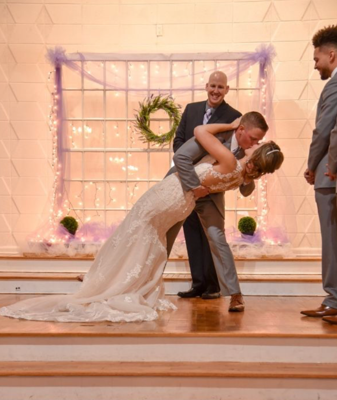 Wedding ceremony: Couple dips, smiling. Groom holds bride, officiant smiles, background decorated.