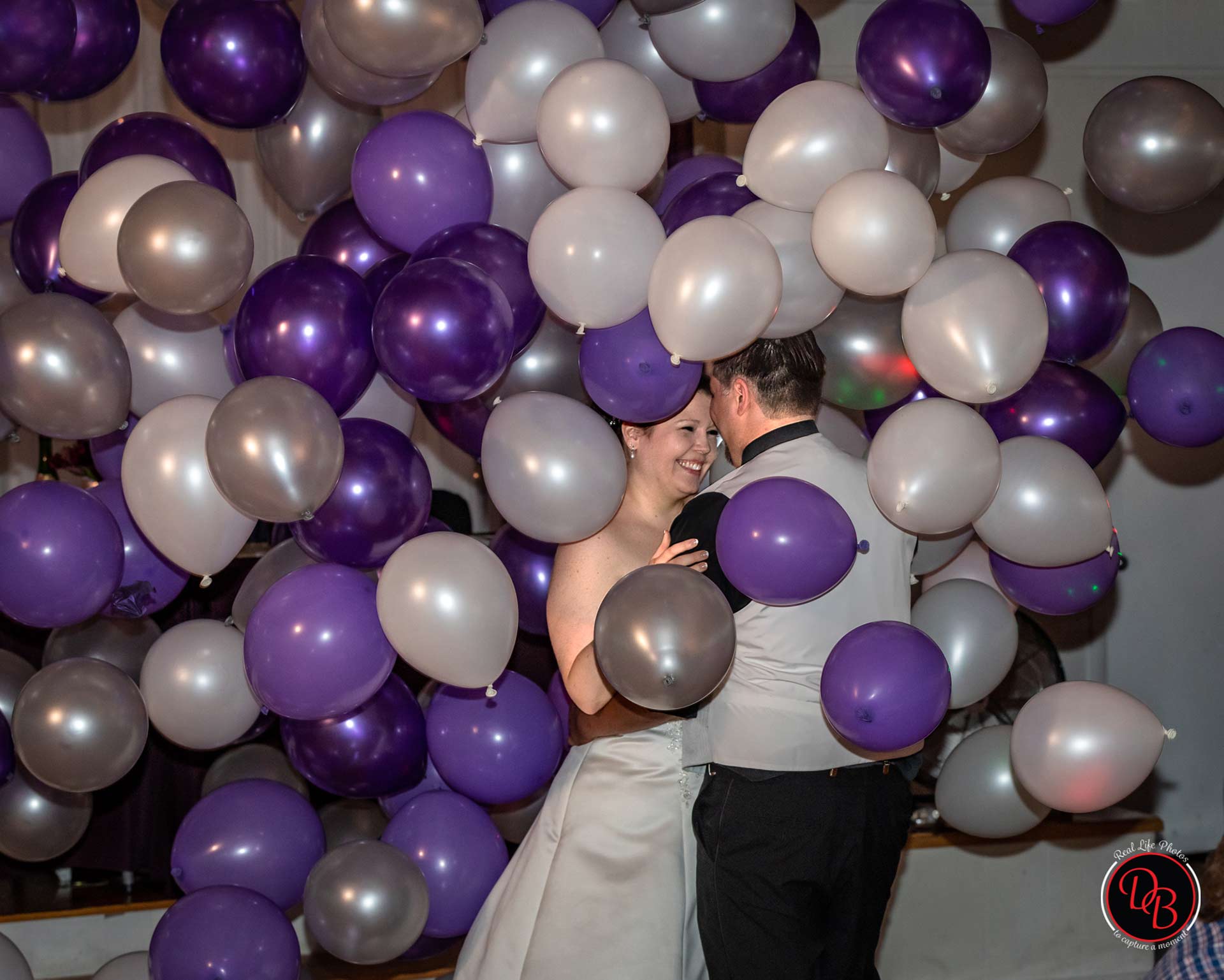 Newlyweds dance surrounded by silver, purple, and white balloons.