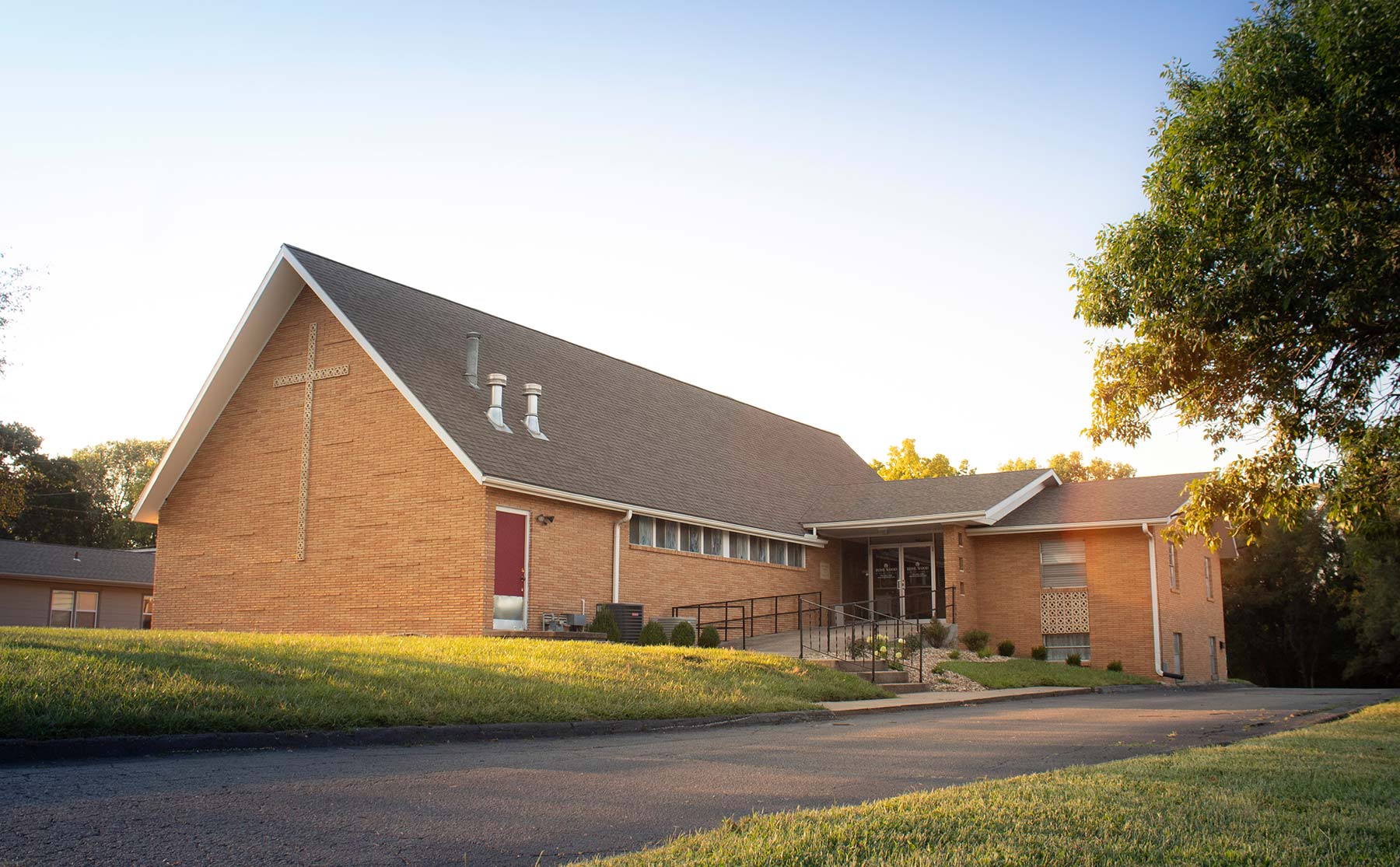 Brick church with a gabled roof and red door on a grassy lawn with a paved road.