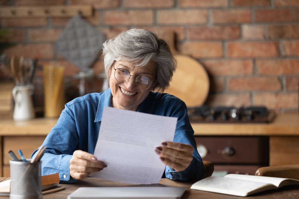 Woman with glasses smiles while reading a letter at a desk in a kitchen.