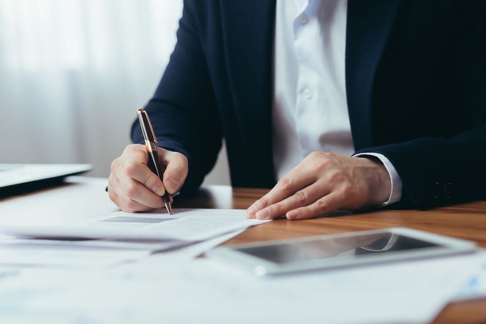 Person in suit writing on paper at a desk, with a pen, phone, and documents visible.