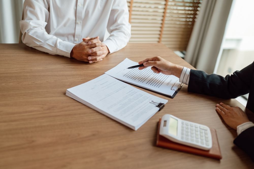 Two people at a desk reviewing documents. One points to a paper; a calculator and notepad sit nearby.