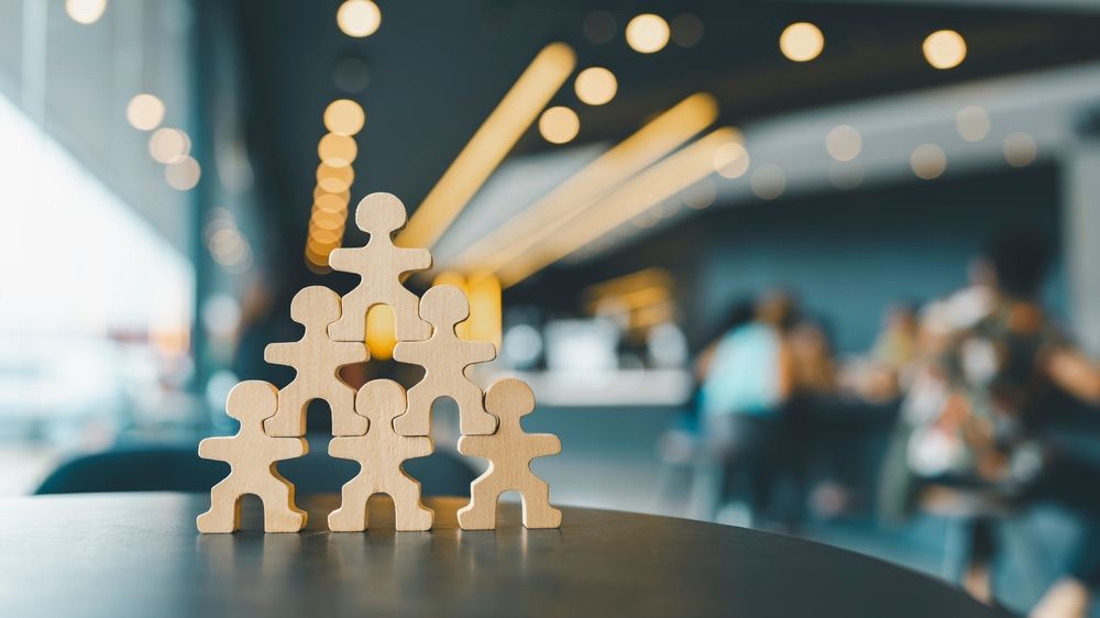Wooden figurines stacked in a pyramid on a table; blurred people in background.