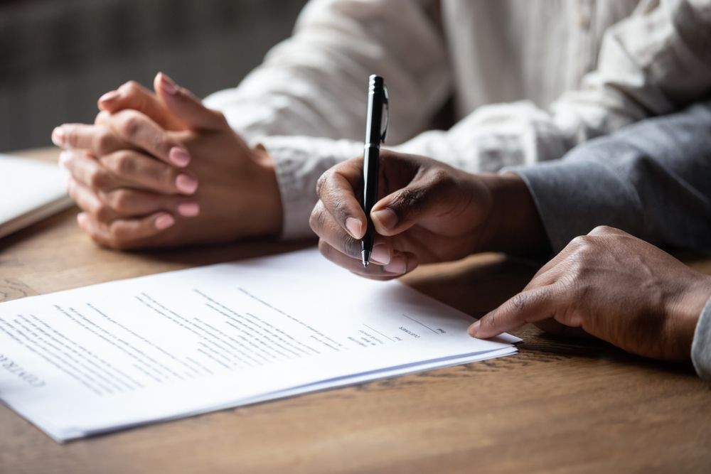 Hands signing a document, one hand holding a pen, another pointing.