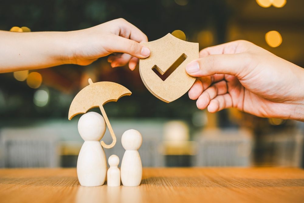 Hands holding shield and umbrella over wooden family figurines on a table.