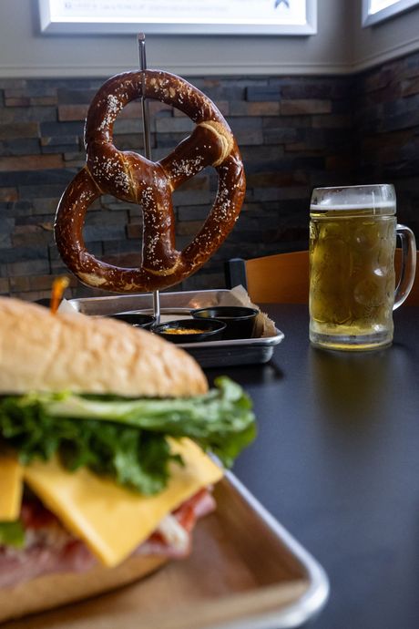 Soft pretzel hanging above a sandwich and a beer mug on a dark table.