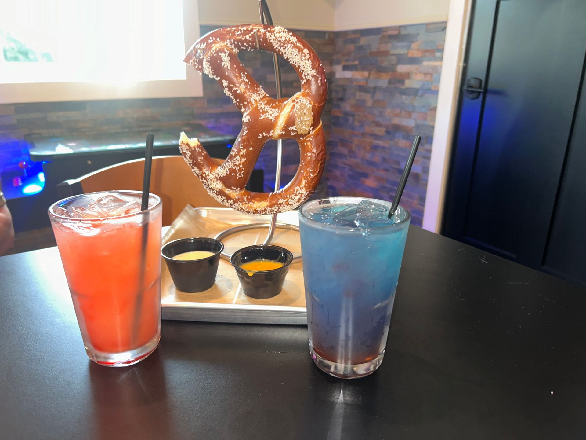 Two colorful drinks with a giant pretzel and dipping sauces on a table.