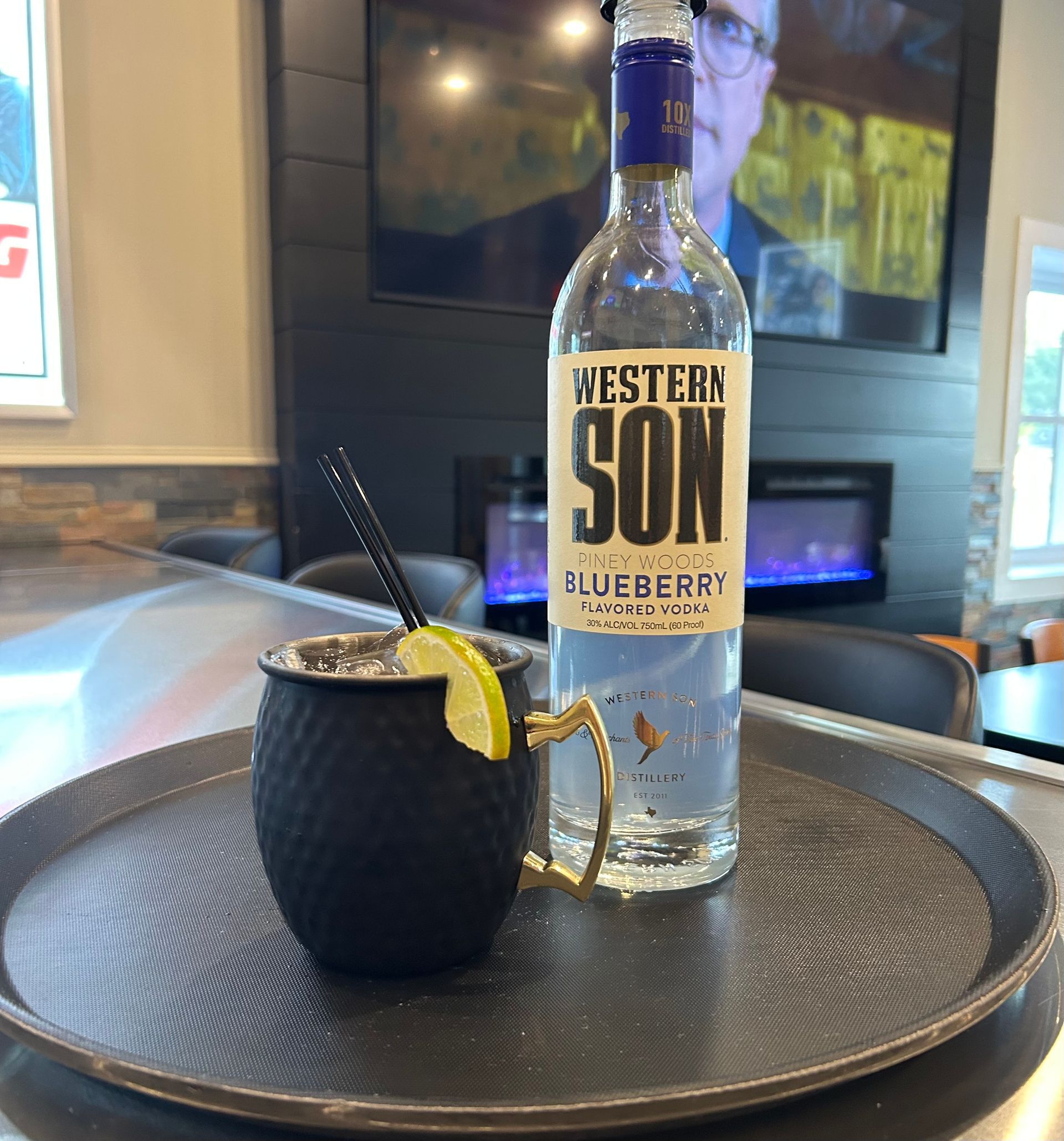Black Moscow mule cocktail and blueberry vodka bottle on a tray, near a fireplace and window.