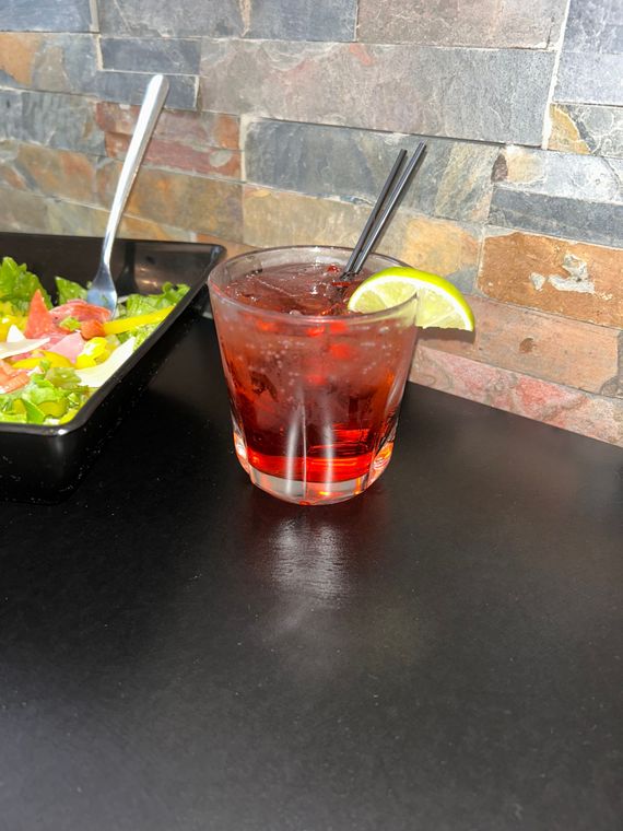 A red cocktail in a glass with a lime wedge and two straws, beside a salad on a black surface.