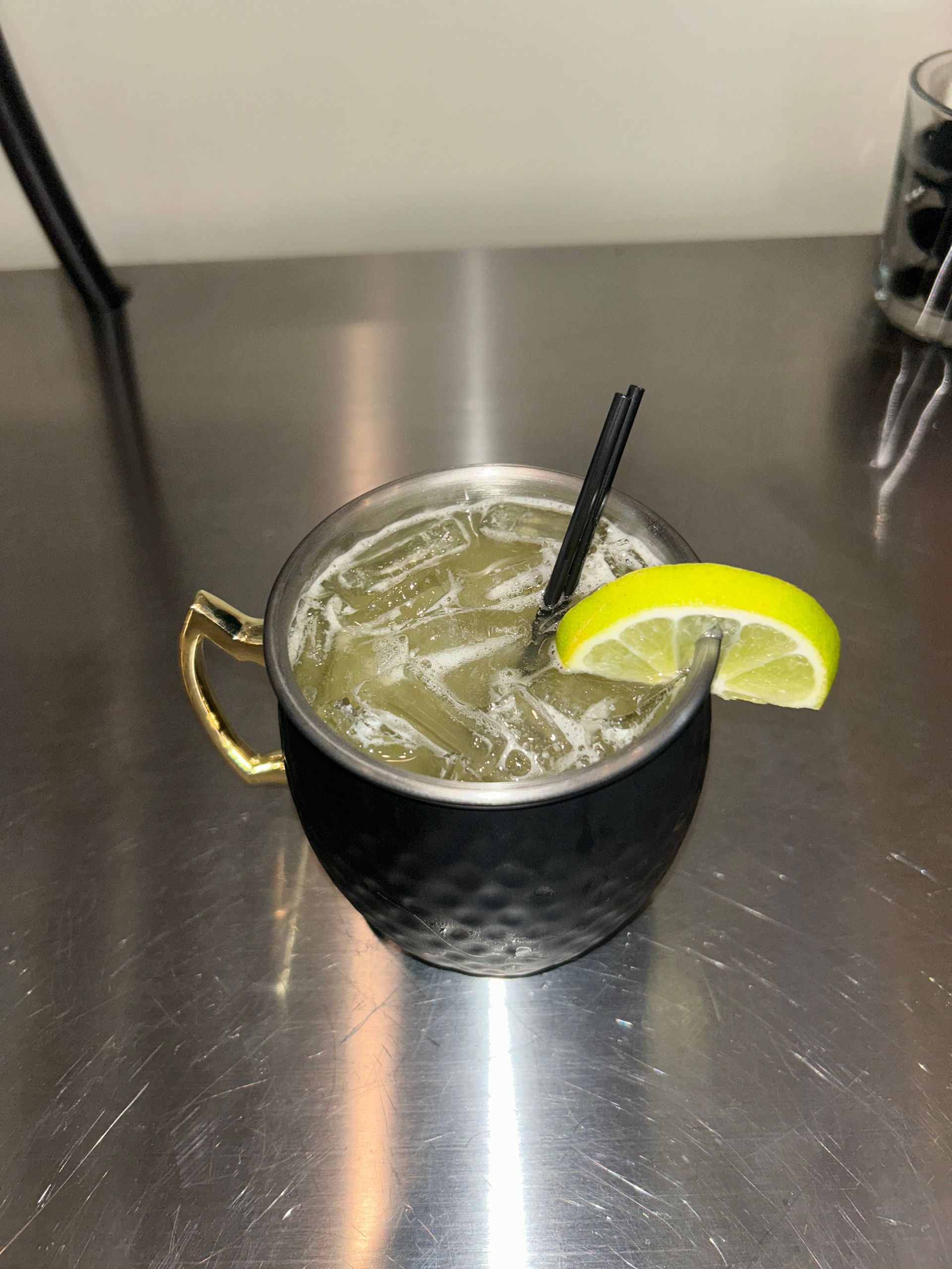 Moscow Mule cocktail in a black mug with gold handle, lime garnish, and black straw on a silver surface.