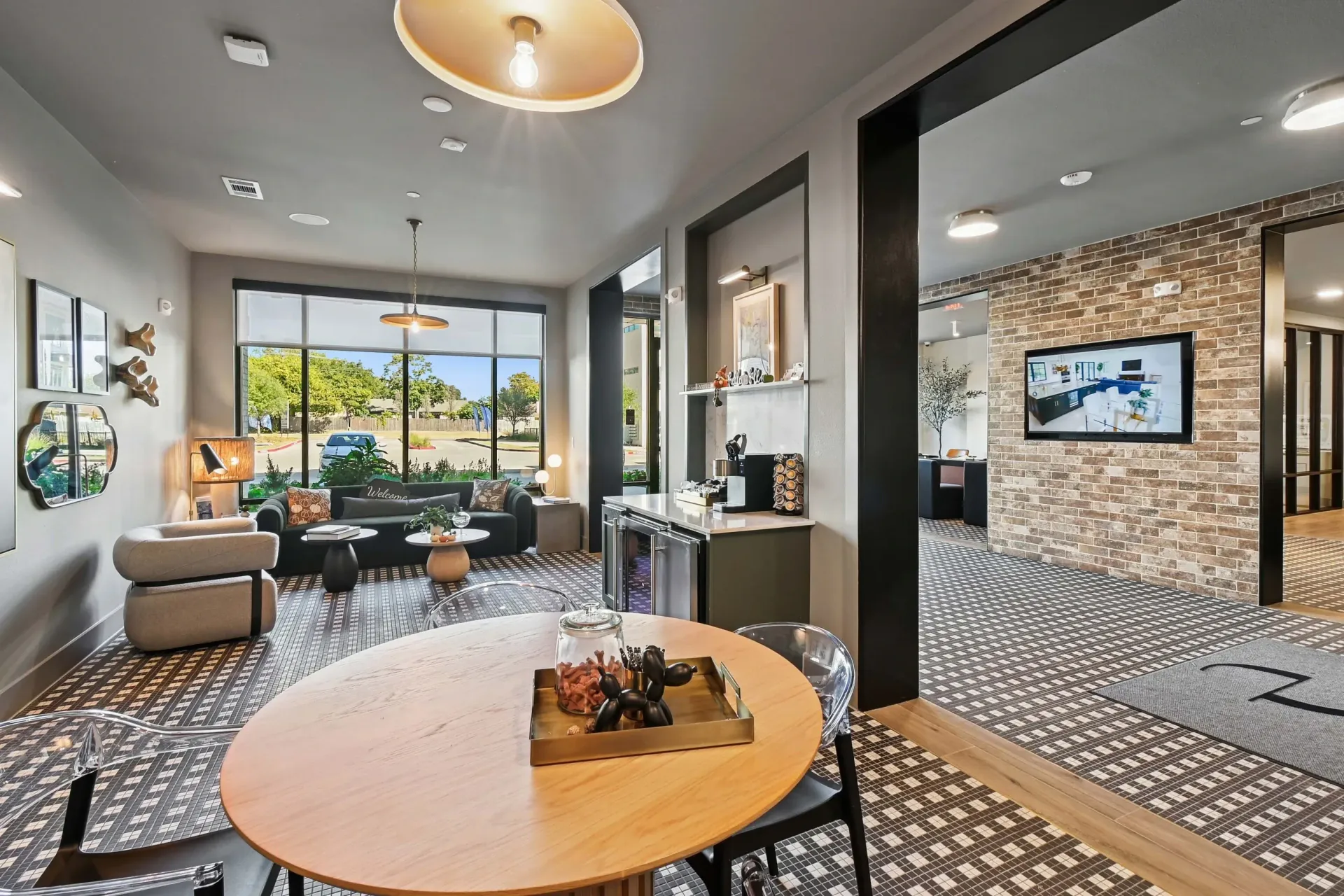 Modern apartment lobby with seating, a round table, large front window, and brick accent wall with a mounted TV at Ennova Highlands apartments in Arlington, TX.