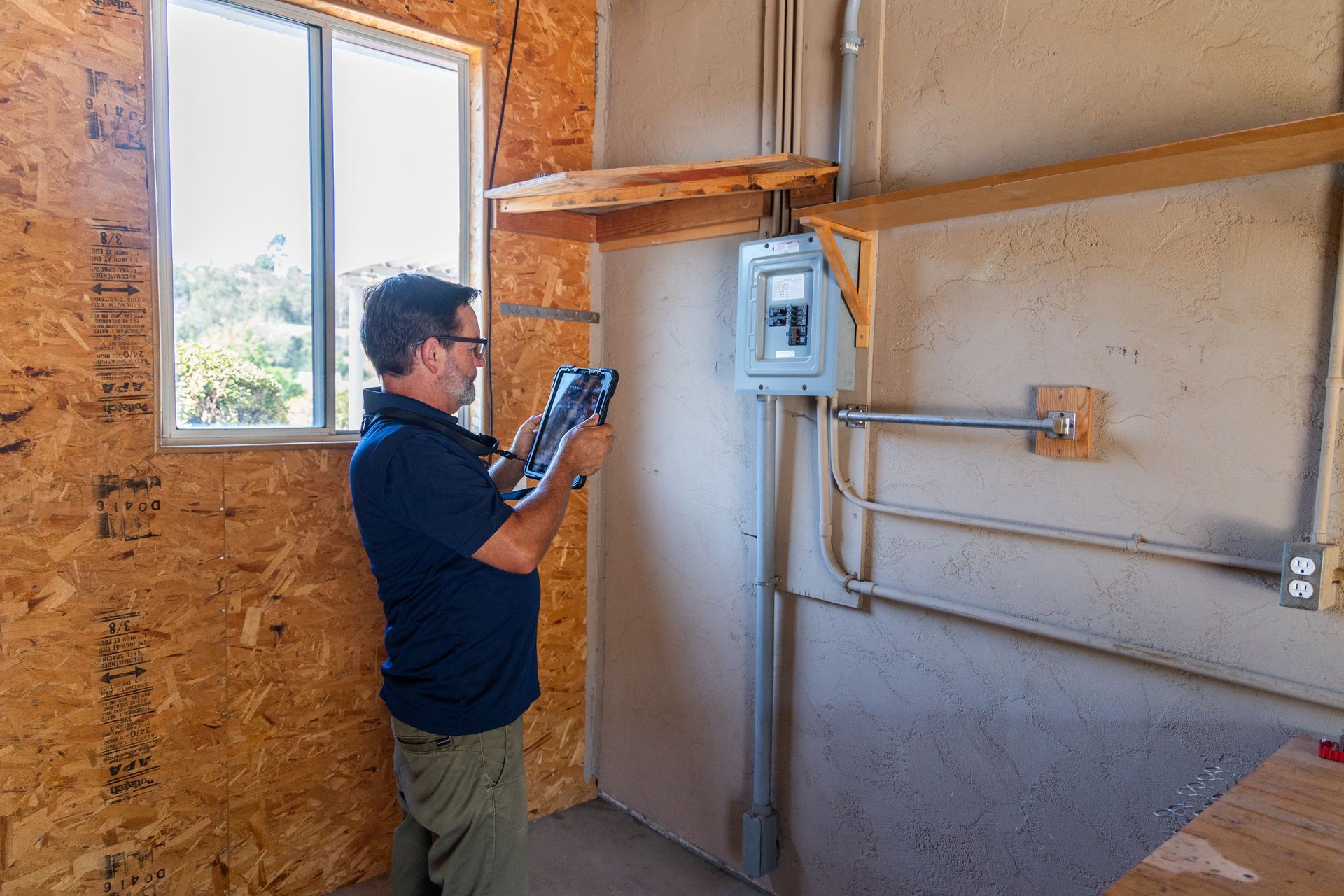 Person inspecting an electrical panel in an unfinished room with exposed wiring and a window. Person inspecting an electrical panel in an unfinished room with exposed wiring and a window.
