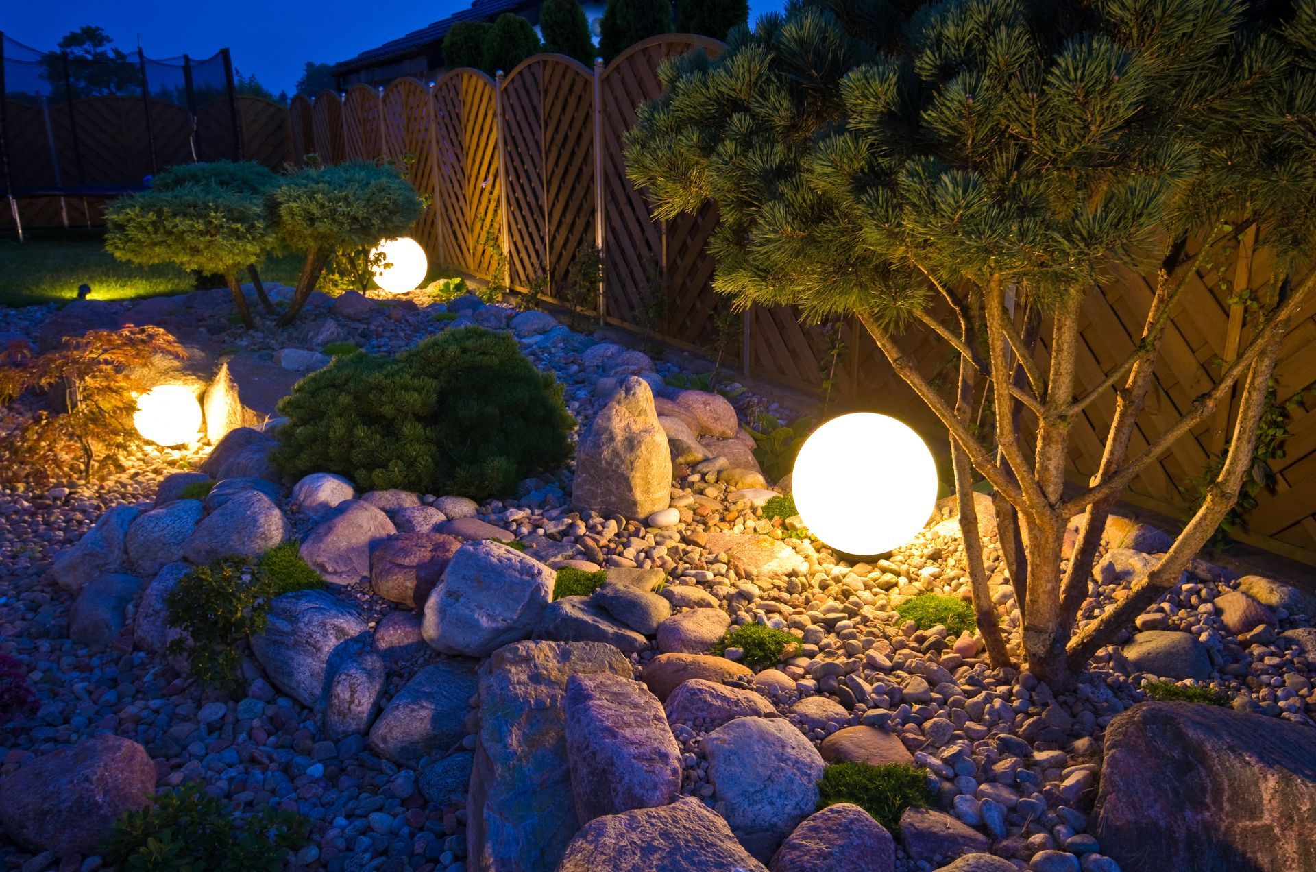 Illuminated garden path with glowing spherical lights, rocks, shrubs, and a wooden fence at dusk