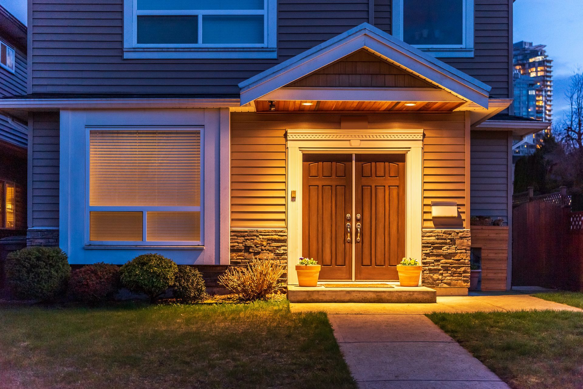 A warmly lit house entryway at dusk with wooden double doors, stone accents, and a small potted plant.