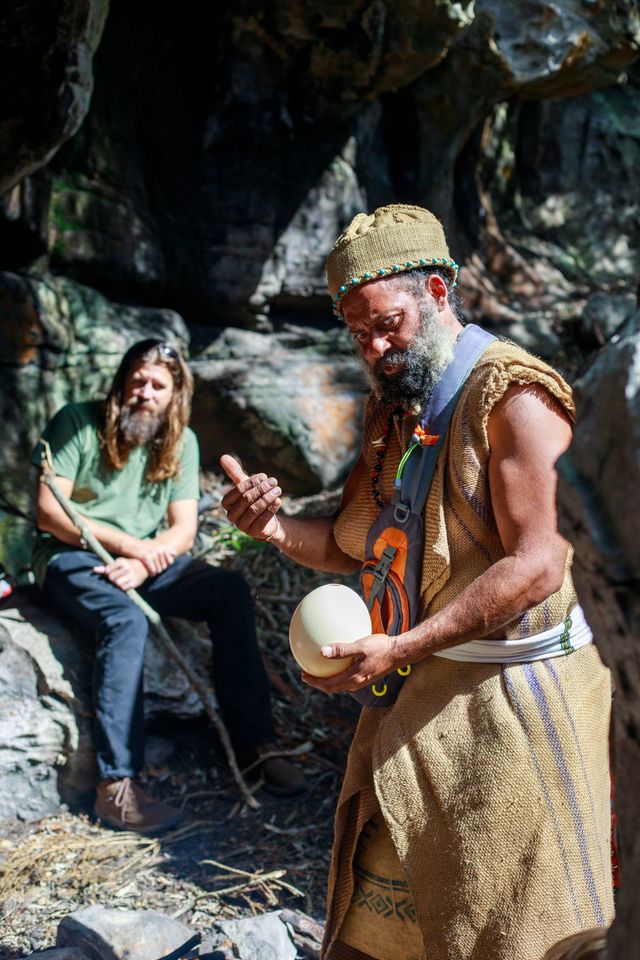A man with a beard is holding an ostrich egg while another man sits on a rock.