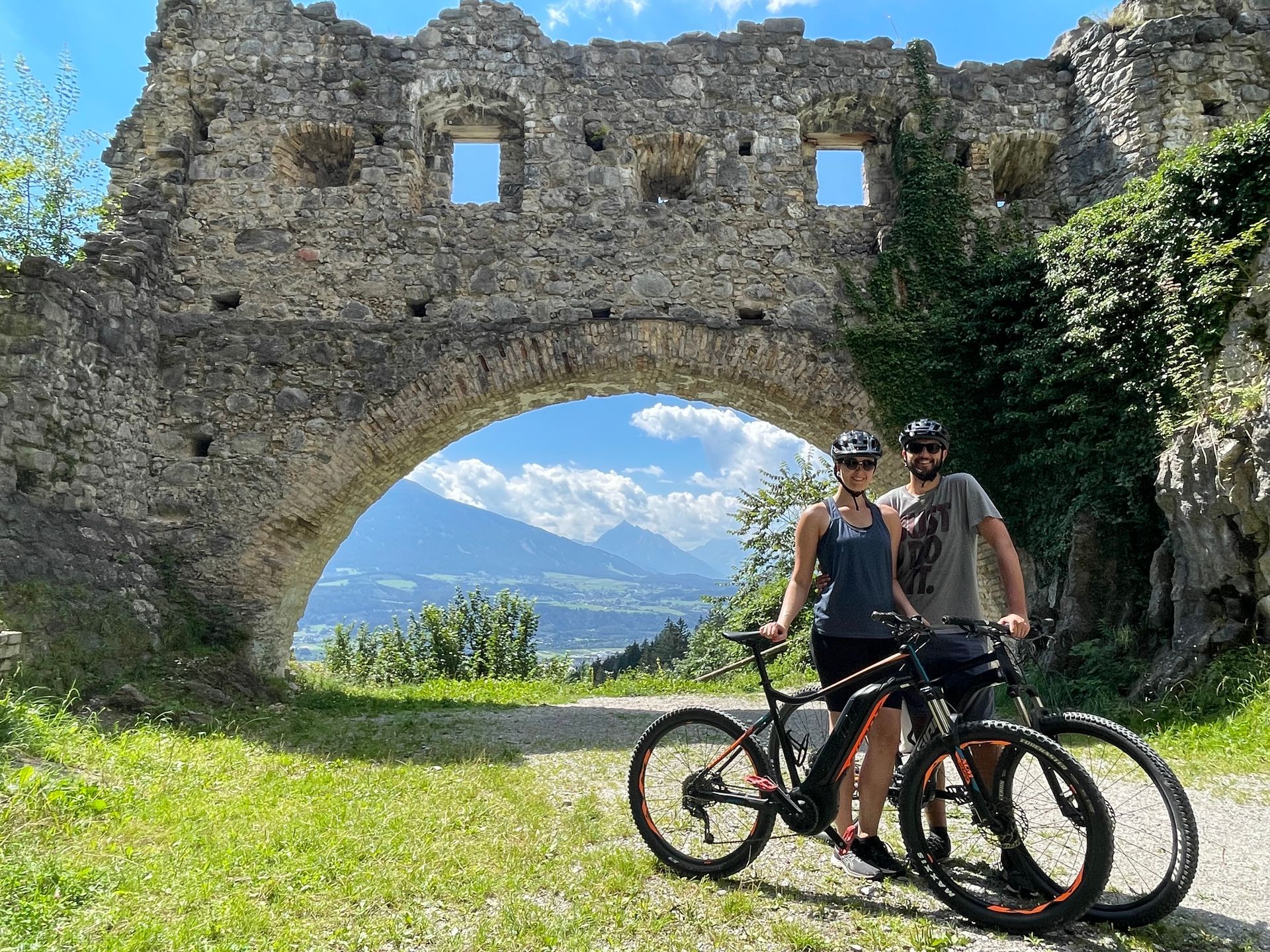 A man and a woman are standing next to their bicycles in front of a stone archway.