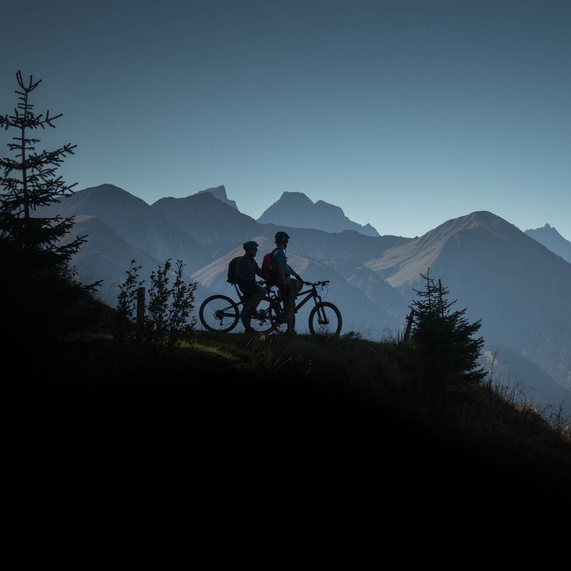 Two people riding bikes on top of a hill with mountains in the background