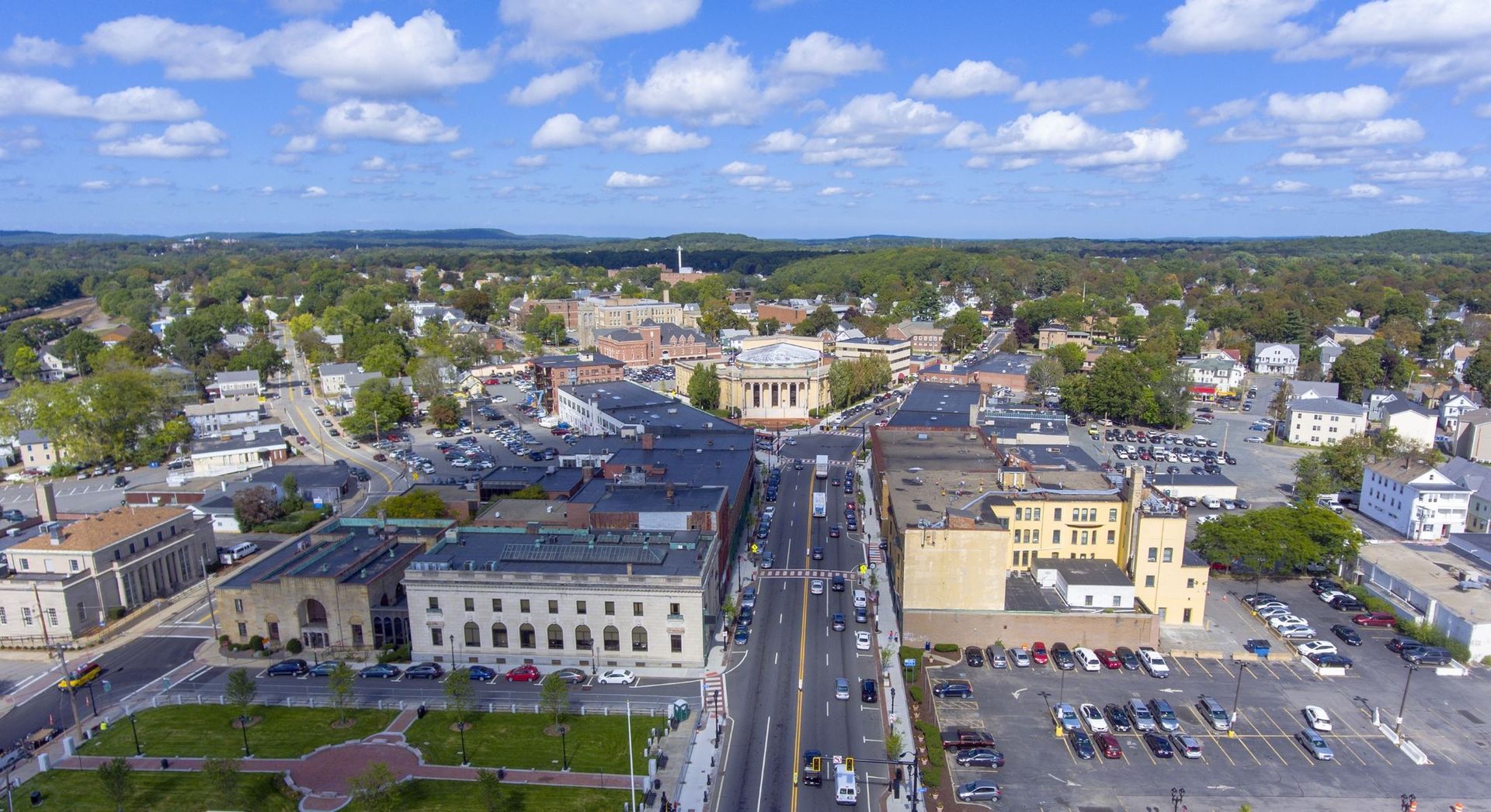 Aerial view of a town square in Framingham, MA with a road, buildings, and parked cars on a sunny day.