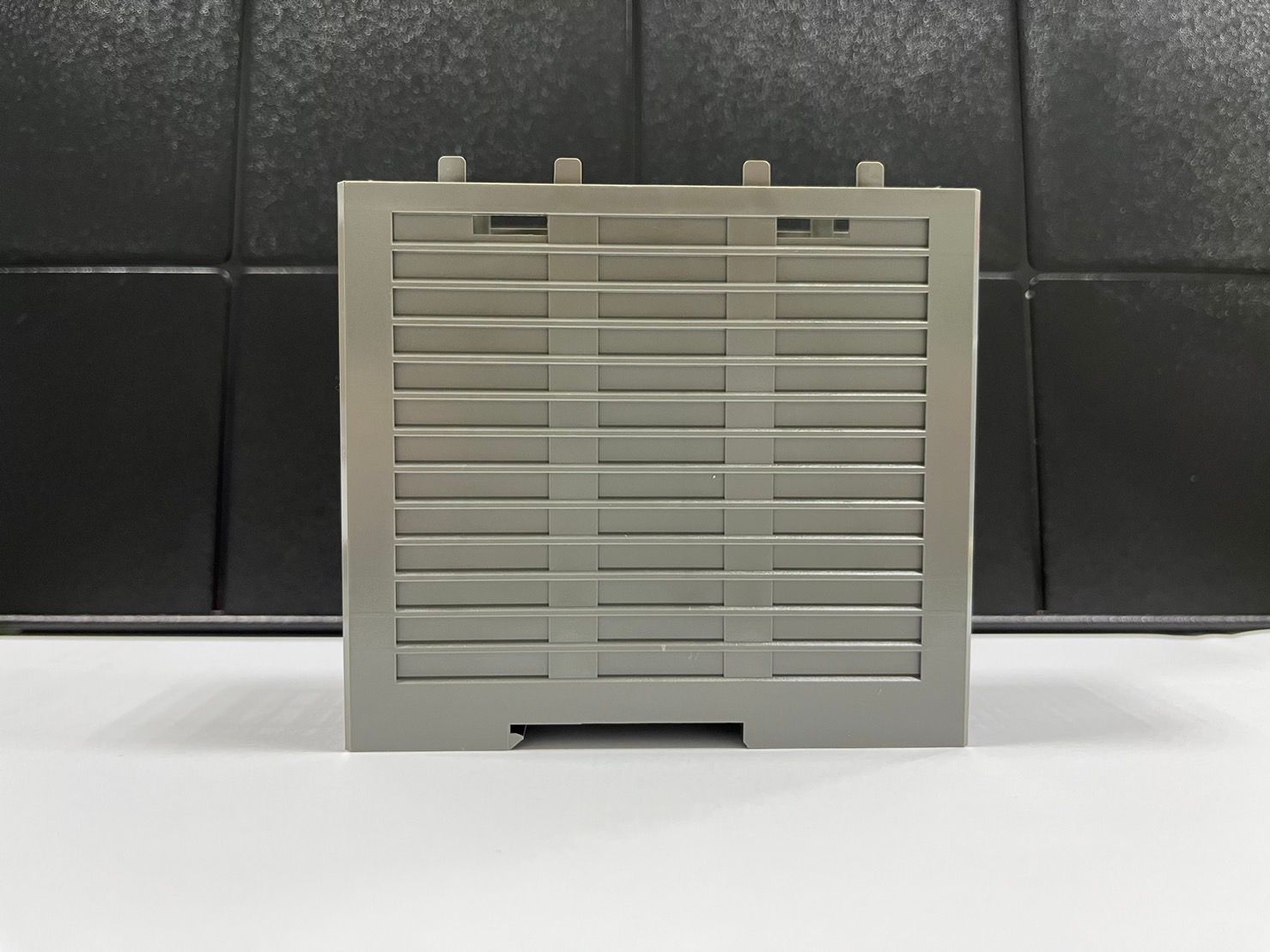 A gray plastic container is sitting on a white surface in front of a black wall.