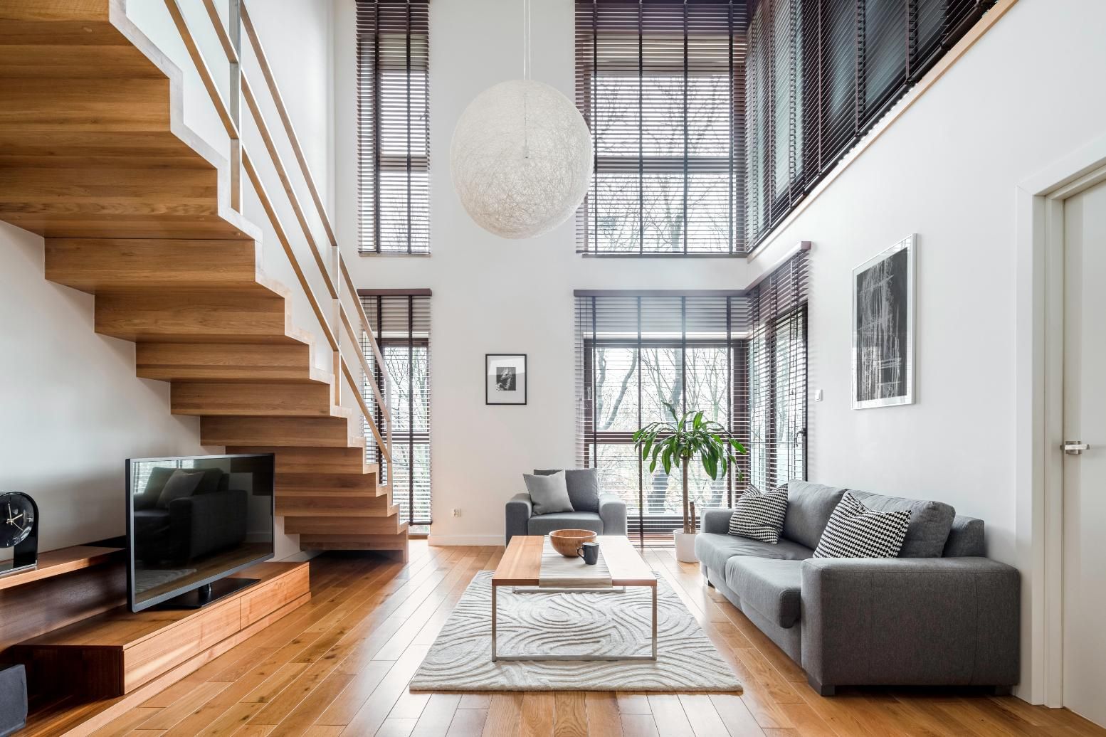 Living room with wooden staircase, gray sofa, coffee table, and large windows with blinds.