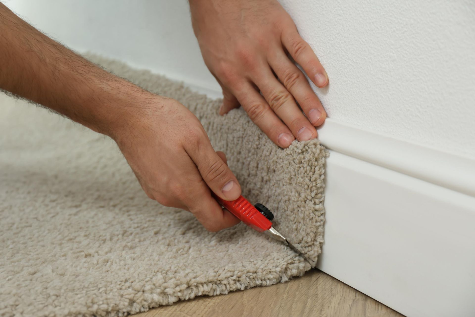 Person using a utility knife to trim beige carpet along a white baseboard in a room.
