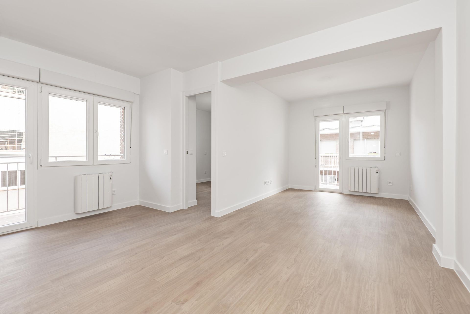 Empty, bright apartment interior with light wood floors, white walls, and two large windows.