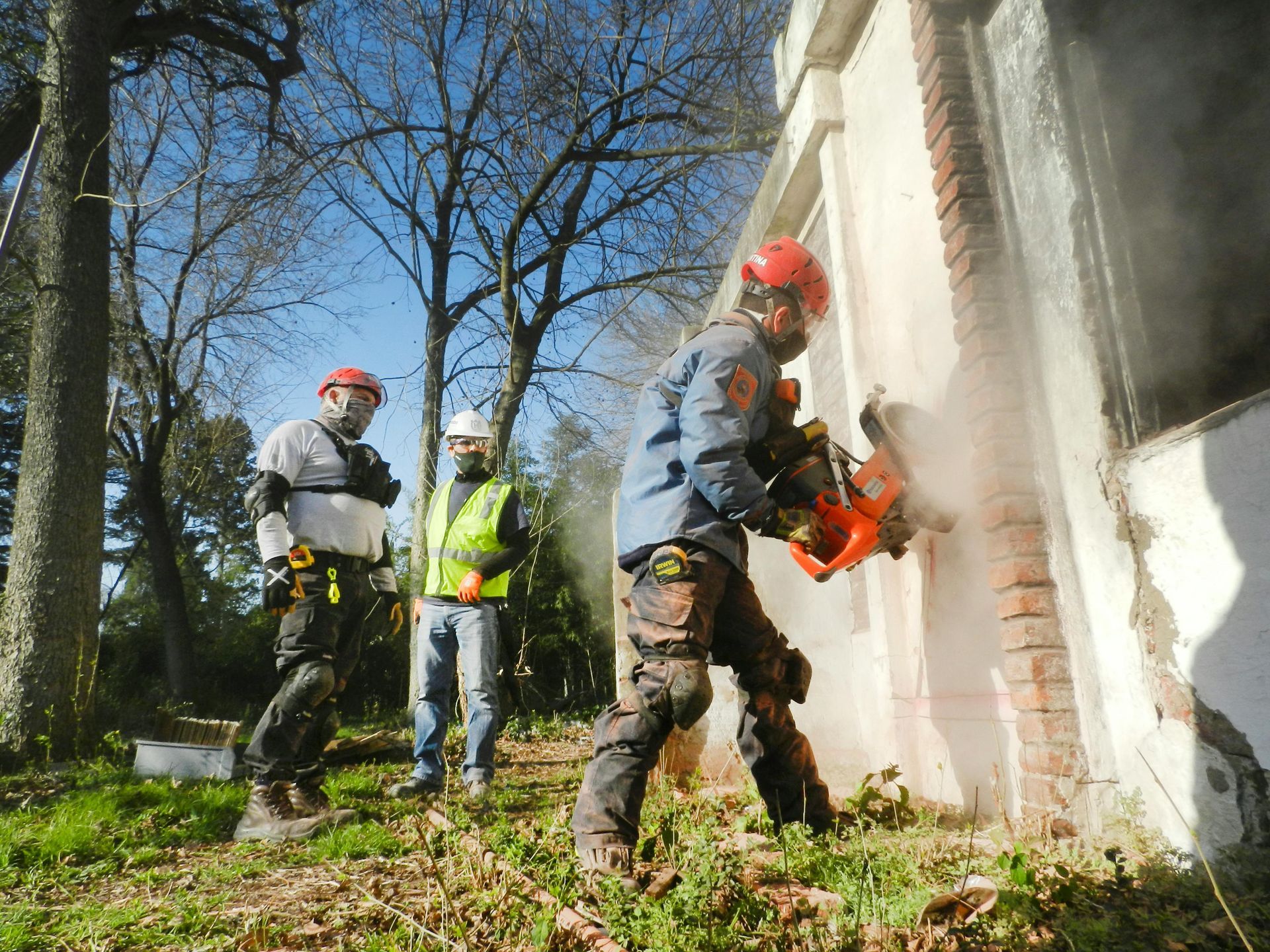 Firefighters cutting a building wall with a saw, wearing helmets and protective gear, outdoors.