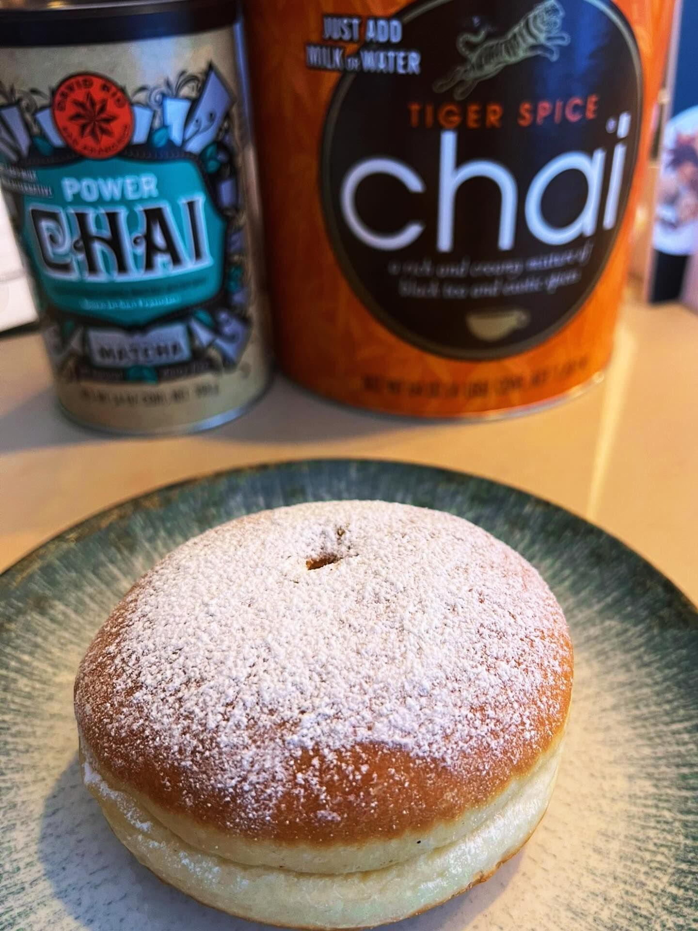 Un pasticcino cosparso di zucchero a velo su un piatto verde, con sullo sfondo lattine di Power Chai e Tiger Spice Chai.