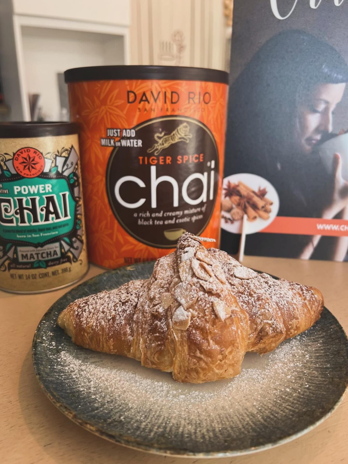 Un croissant fresco cosparso di mandorle in polvere, adagiato su un piatto grigio, si trova davanti a una scatola di tè David Rio Tiger Spice Chai e Power Chai.