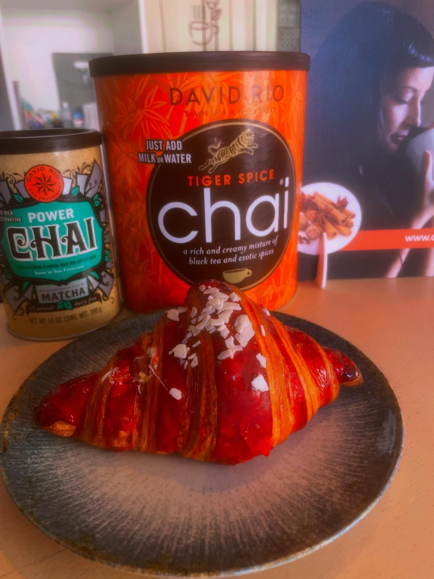 Un croissant glassato di rosso è adagiato su un piatto davanti a una bottiglia di tè chai Tiger Spice e a una lattina di tè matcha Power Chai.