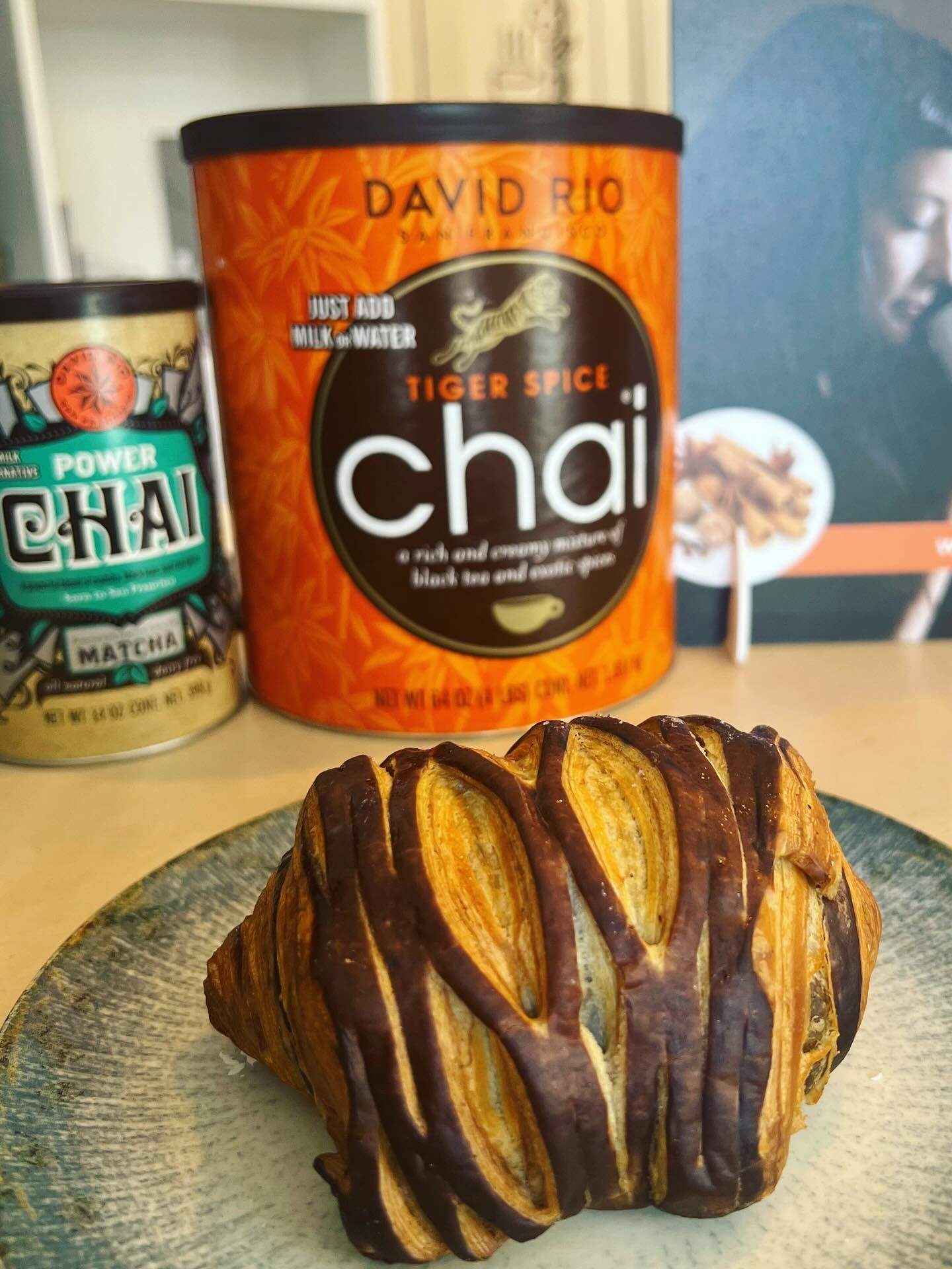 Un croissant ricoperto di cioccolato su un piatto decorato, con contenitori di David Rio Tiger Spice e Power Matcha chai mix.