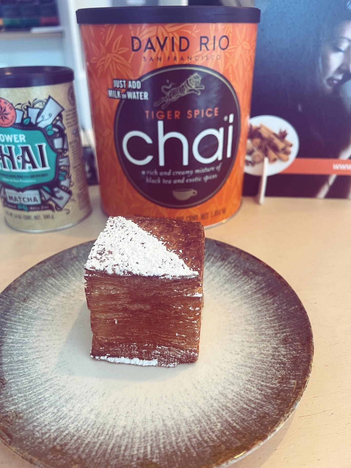 Un pasticcino a forma di cubo ricoperto di zucchero a velo su un piatto dalla superficie ruvida, con sullo sfondo delle lattine di tè chai David Rio Tiger Spice.