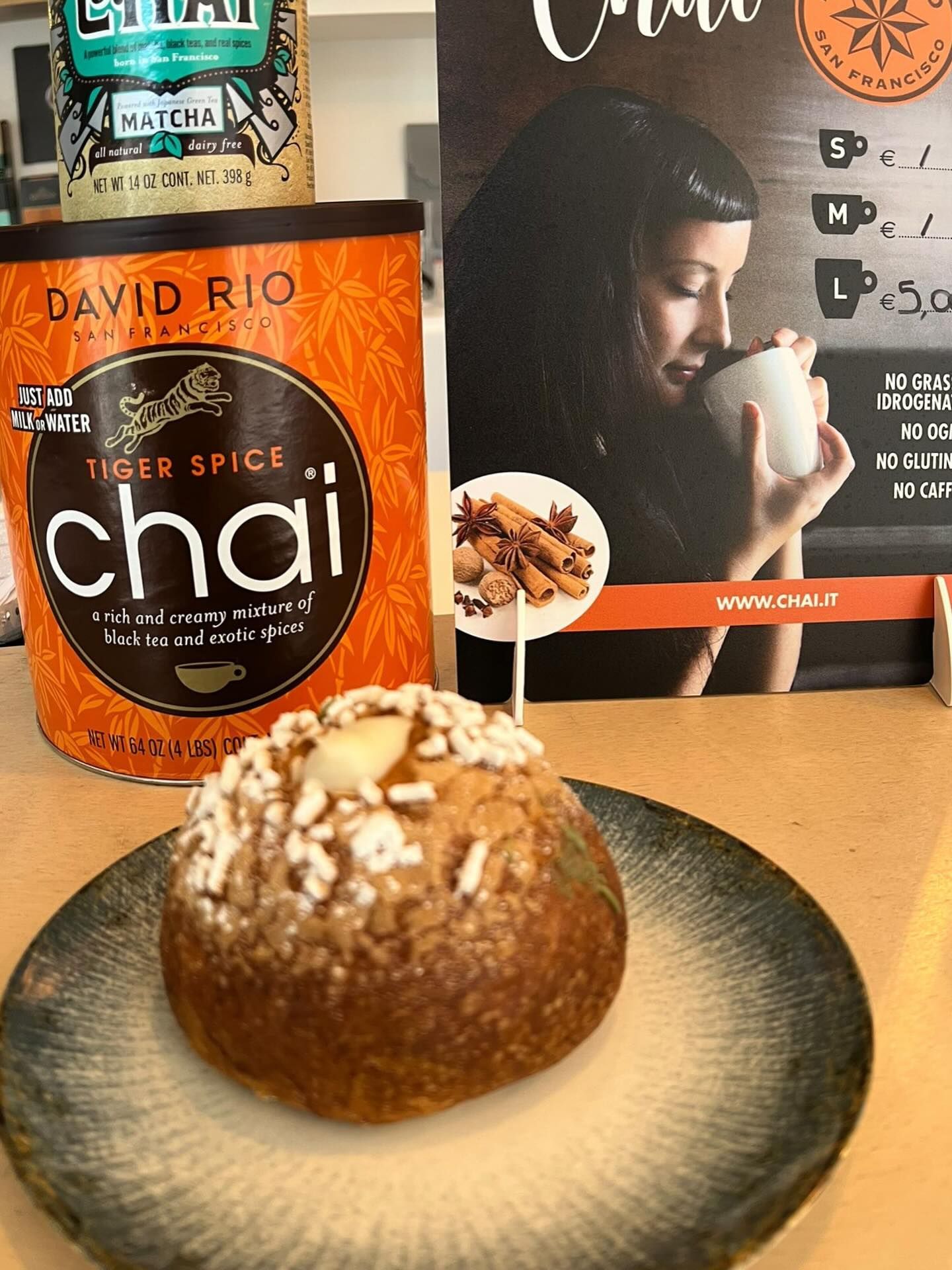 Un pasticcino ricoperto di zucchero è adagiato su un piatto davanti a una lattina di tè chai David Rio Tiger Spice e a un espositore con il menù promozionale.