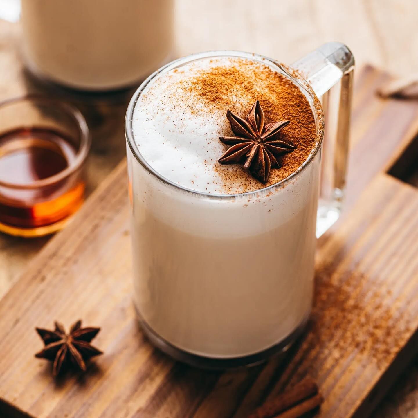 Una tazza di latte macchiato in vetro, guarnita con schiuma, cannella e anice stellato, poggiata su un tagliere di legno con spezie e sciroppo nelle vicinanze.