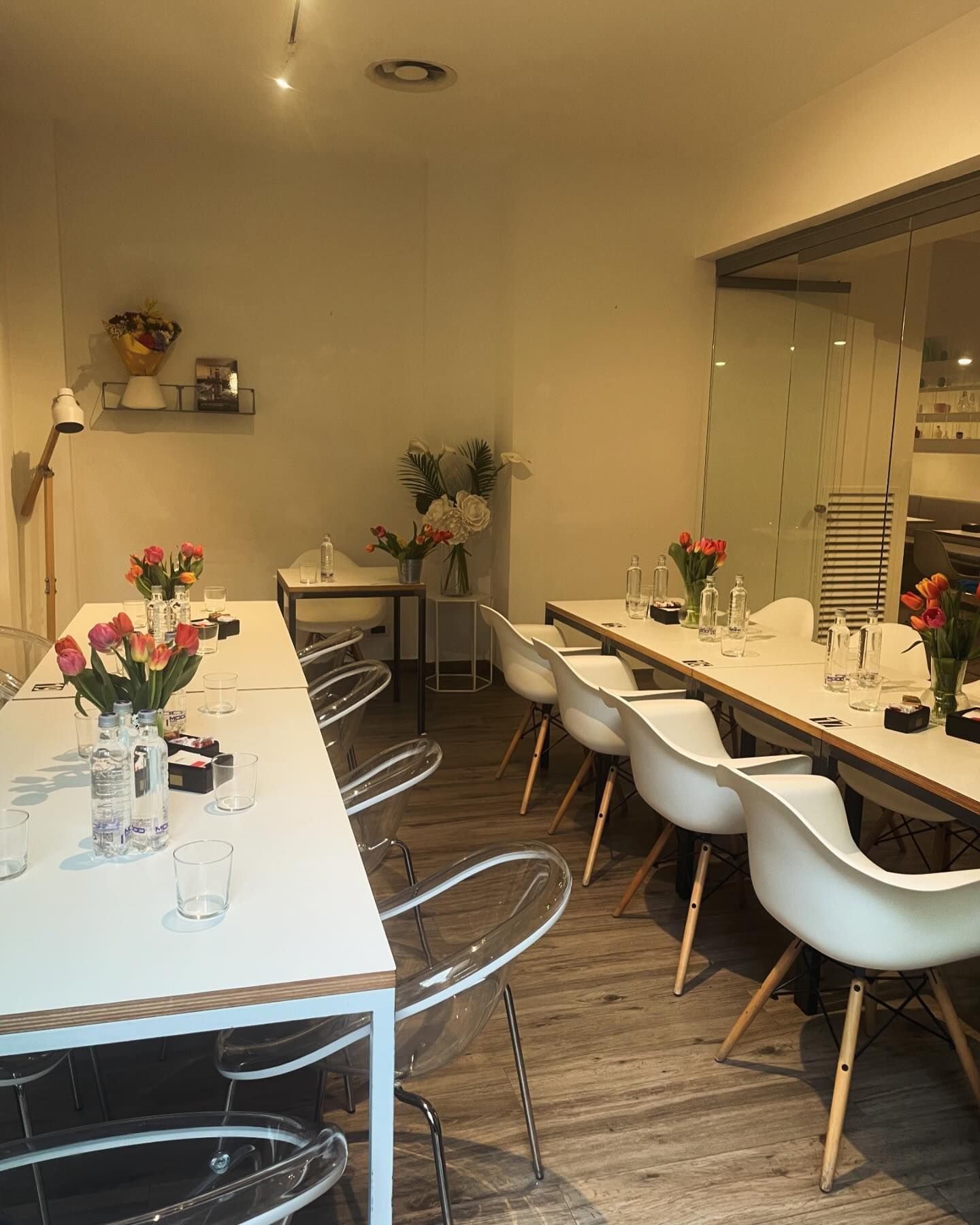 Una stanza luminosa con due lunghi tavoli bianchi, sedie trasparenti, sedie bianche e vasi di fiori, allestita per un incontro.