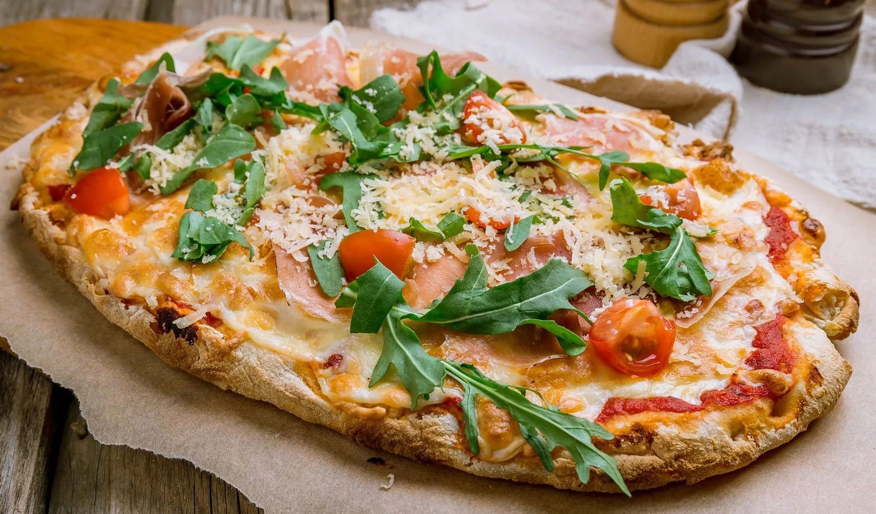 Pizza su pane piatto condita con formaggio fuso, salsa di pomodoro, pomodorini, prosciutto crudo e rucola fresca, servita su carta da forno.