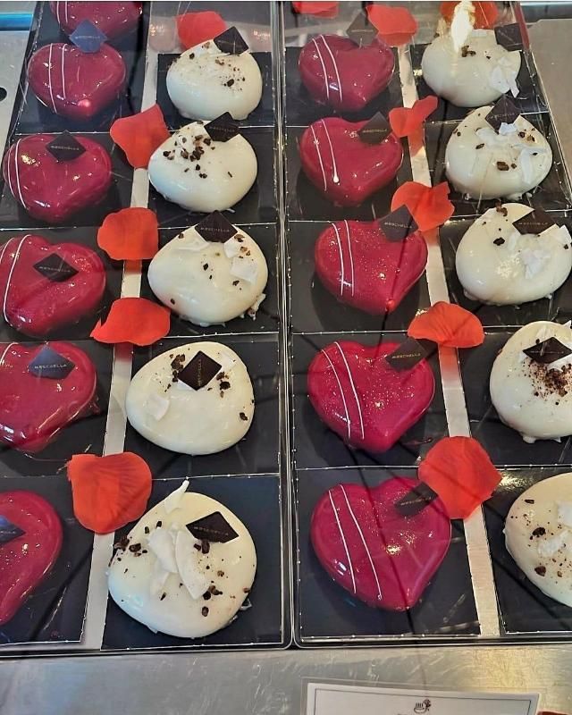 Una griglia di tortine a forma di cuore con glassa lucida rossa e bianca, decorate con piccoli quadratini di cioccolato fondente e petali di rosa.