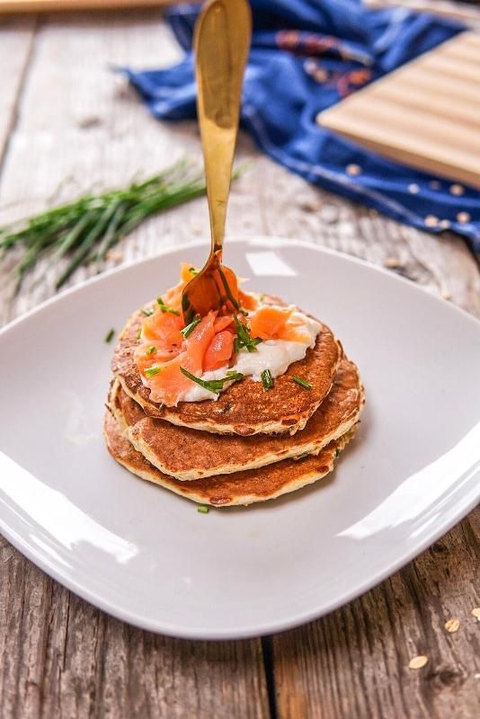 Una pila di tre pancake dorati, guarniti con formaggio cremoso, salmone affumicato ed erba cipollina fresca, serviti su un piatto bianco.