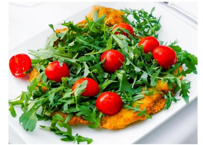Una cotoletta impanata guarnita con rucola fresca e pomodorini rossi, servita su un piatto rettangolare bianco.