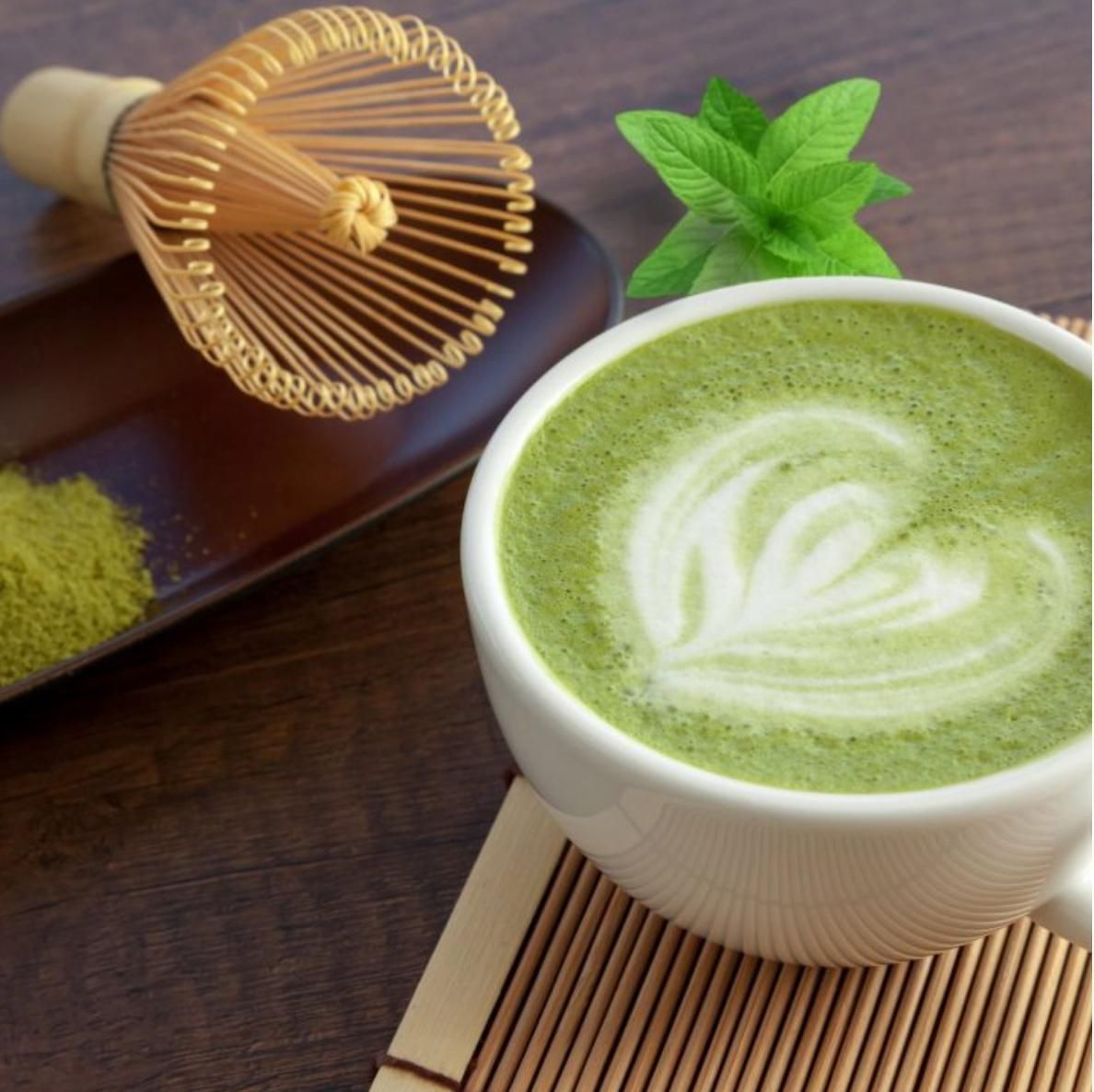 Una tazza di spumoso matcha latte con decorazioni artistiche, accanto a una frusta di bambù, polvere verde e foglie di menta su un tavolo di legno.