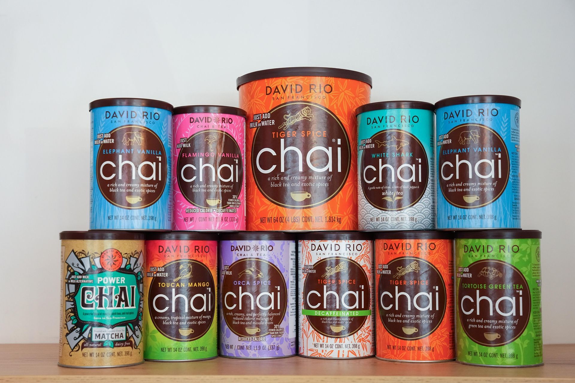 Una collezione di undici barattoli di tè chai in polvere David Rio, di vari colori e dimensioni, disposti su una superficie di legno.