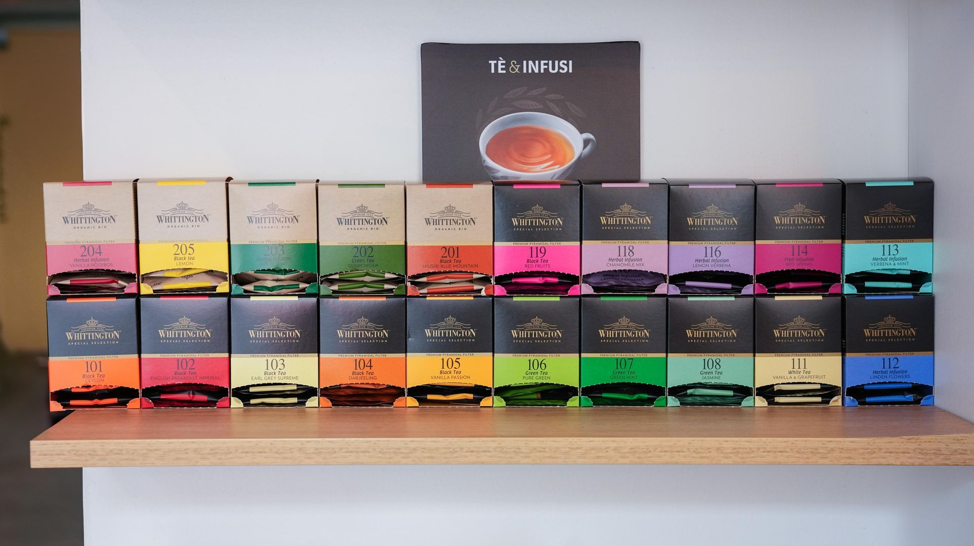 Su uno scaffale di legno, addossato a una parete bianca, sono disposte due file di scatole di tè colorate, ognuna con un'etichetta che raffigura una tazza di tè.