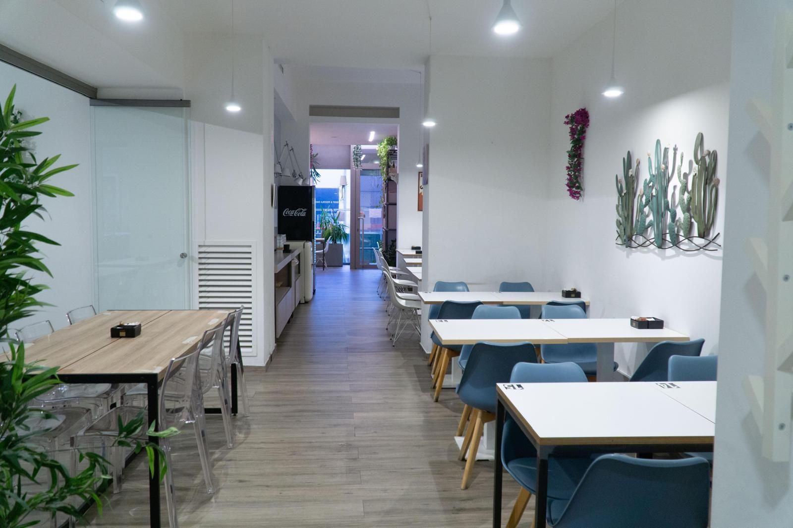 Vista interna di una moderna sala da pranzo con tavoli in legno chiaro, sedie blu e quadri raffiguranti cactus su pareti bianche.