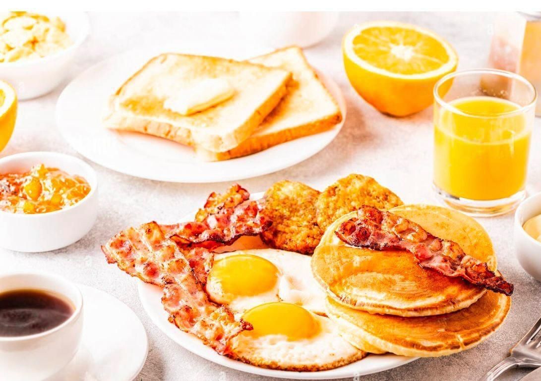 Una colazione completa con uova, pancetta, pancake, pane tostato, succo d'arancia e caffè su una superficie di colore chiaro.