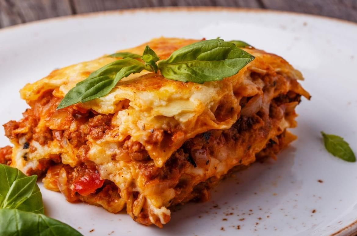 Una fetta di lasagna dorata, guarnita con foglie di basilico fresco, su un piatto bianco.
